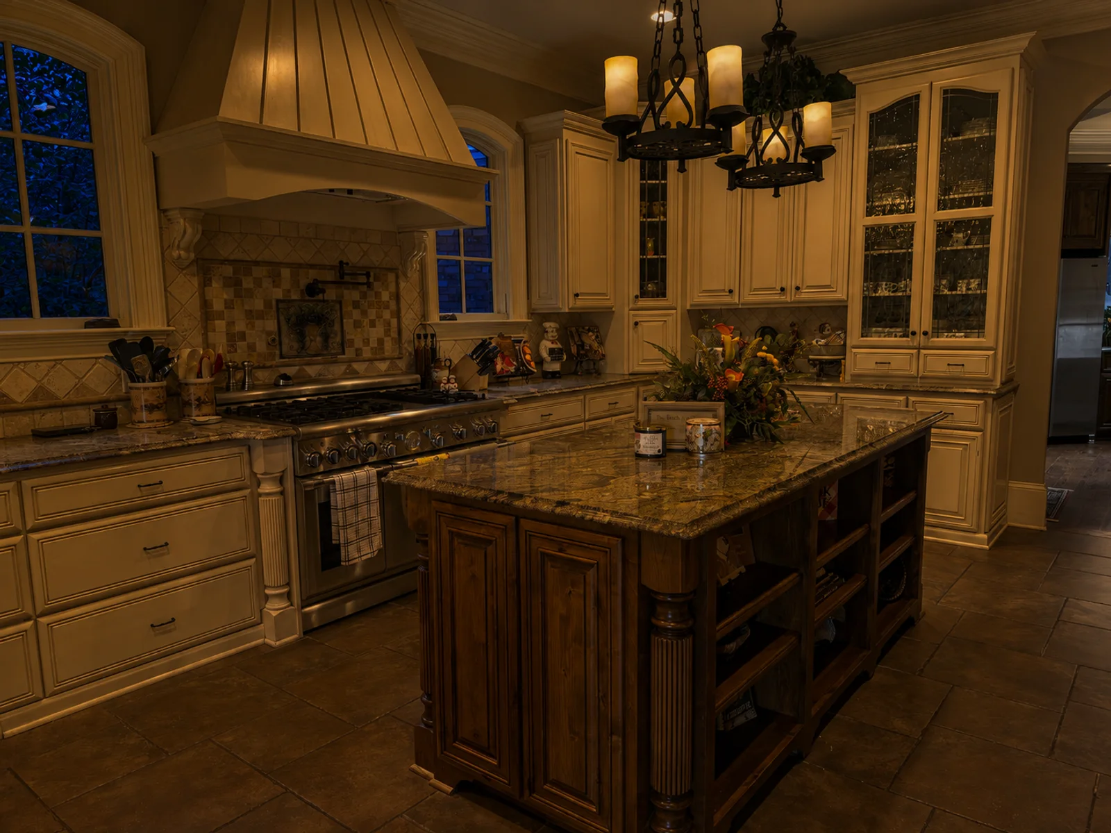 AI evening lighting interpretation of the same view of an ivory kitchen with a timber island and two chandeliers